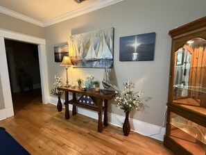 A room with a wooden floor, a console table with decorative items, a lamp, a painting, and a cabinet. - Anchored in Paradise, Walk to Beach & Island View Casino (Gulfport)