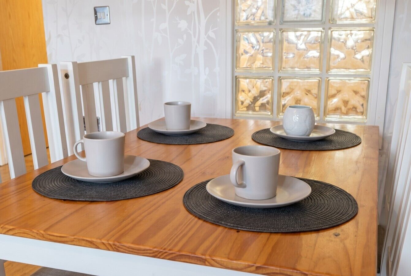 A wooden dining table with four white cups and saucers, black placemats, and white chairs.