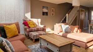 A living room with a sofa, armchair, and ottoman, a wooden coffee table, and a staircase leading to another room.
