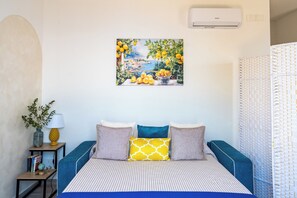 A bedroom with a bed, pillows, a bedside table with a lamp, a painting, and a window with a view. - Santa Rosa Relais | Amalfi Coast (Conca dei Marini)
