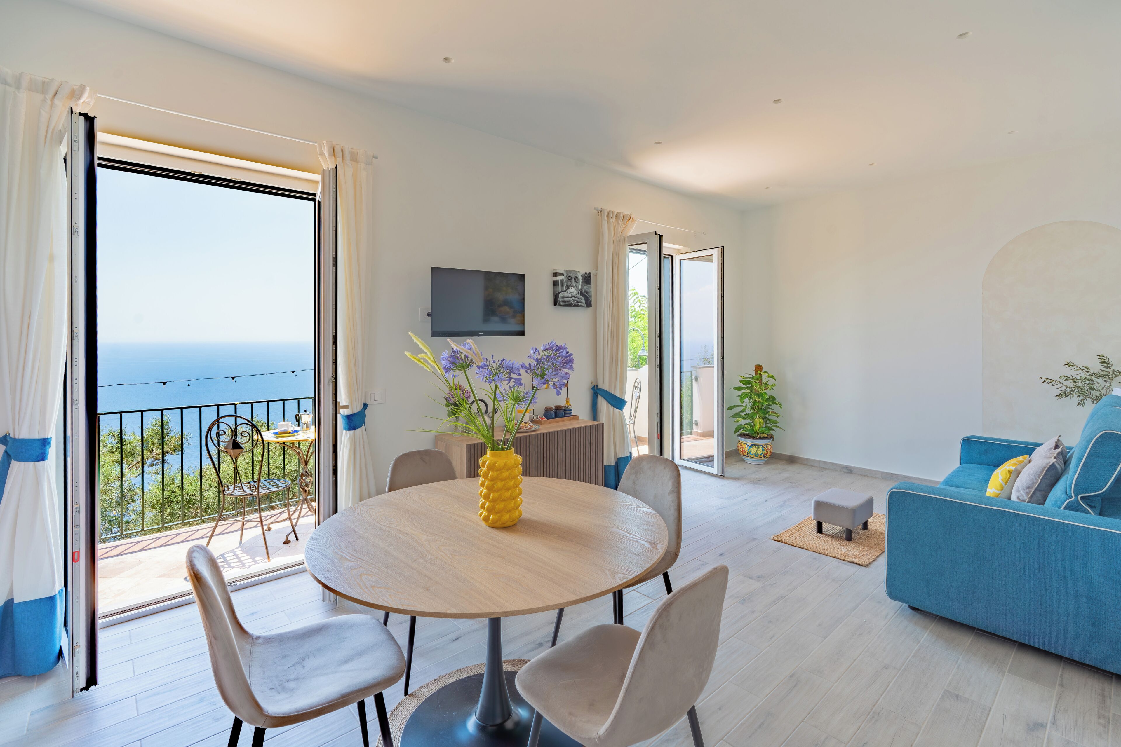 Une pièce avec un balcon, une table à manger ronde entourée de chaises, un canapé et un téléviseur fixé au mur.