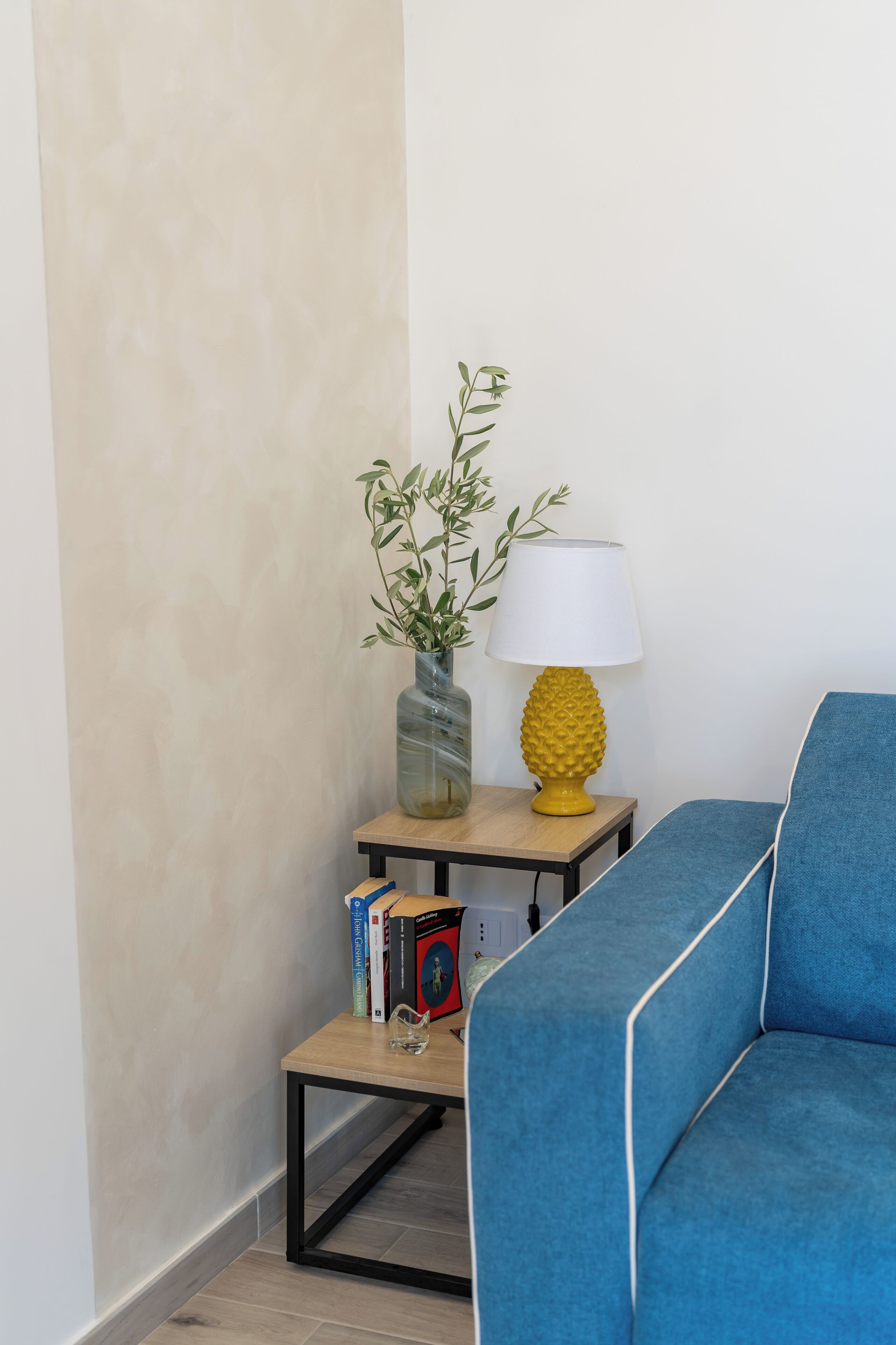 A corner of a room with a blue sofa, a small table with a lamp, a vase with a plant, and a few books.