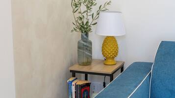 A corner of a room with a blue sofa, a small table with a lamp, a vase with a plant, and a few books.
