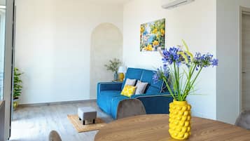 Una sala moderna con un sofá azul, una mesa de comedor de madera y un jarrón amarillo con forma de piña lleno de flores.