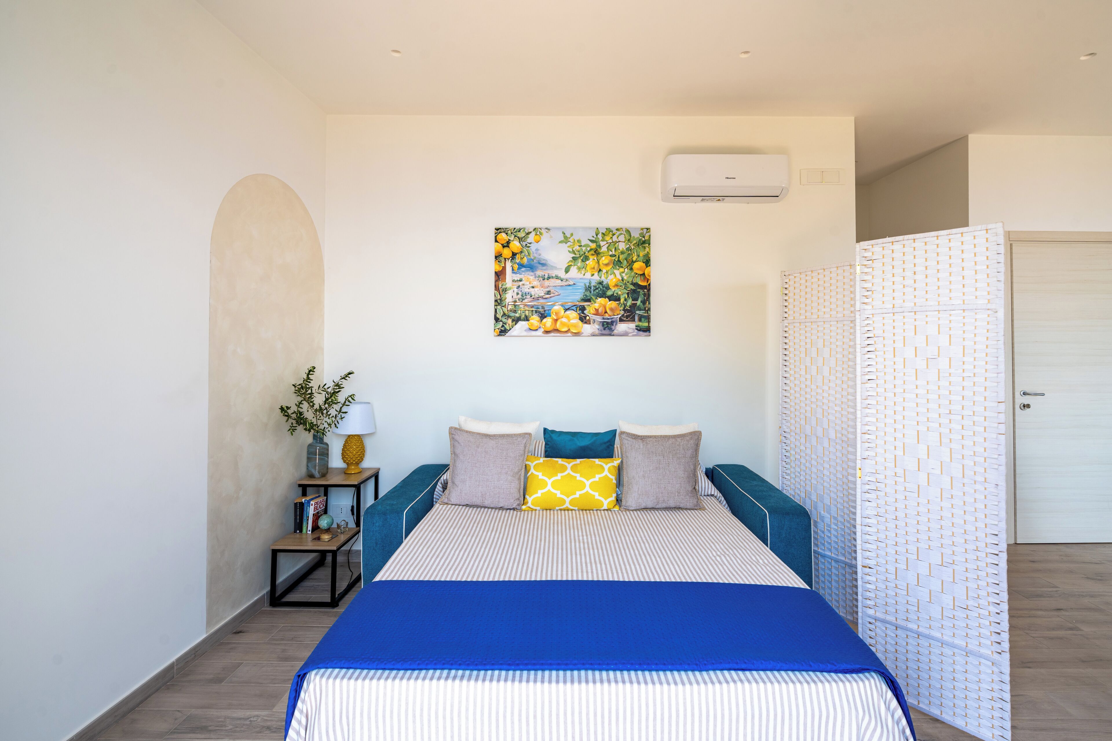 A bedroom with a bed, a painting, a small table with a plant, and a door.