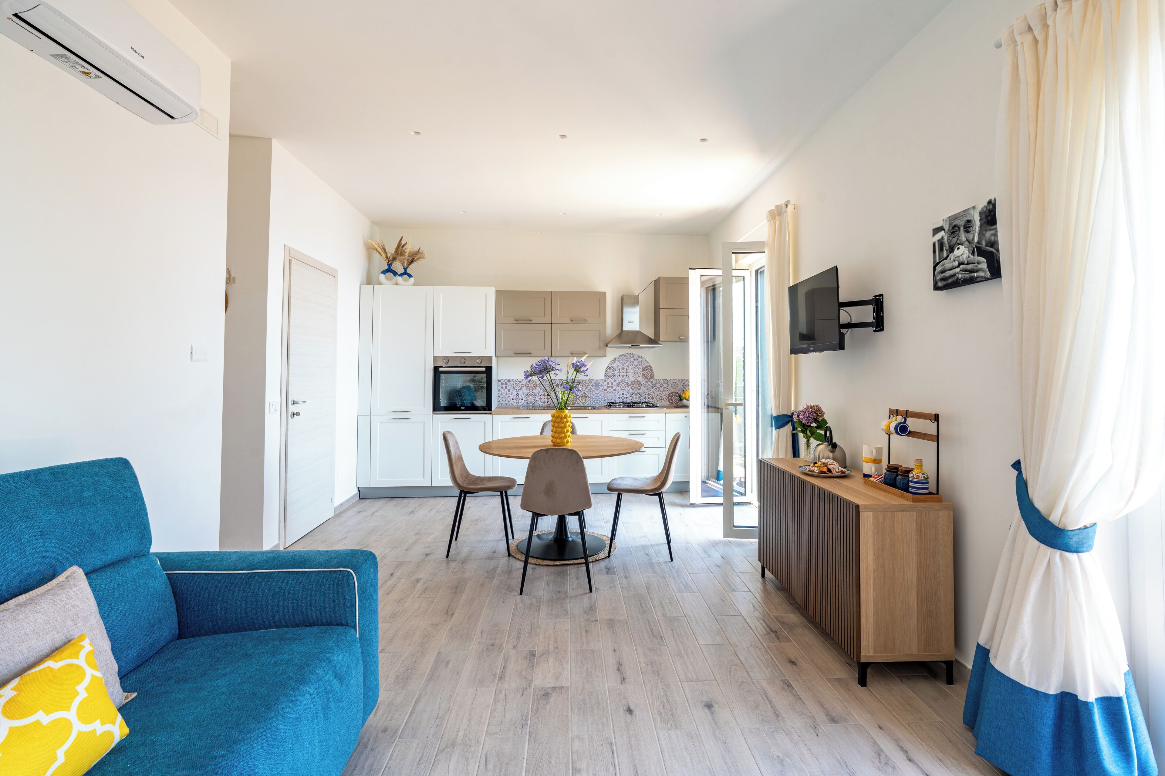 A modern living room with a blue sofa, a dining area with a round table, and a kitchen with white cabinets and a built-in oven.
