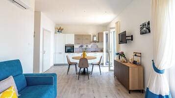 A modern living room with a blue sofa, a dining area with a round table, and a kitchen with white cabinets and a built-in oven.