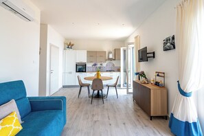 Una sala de estar moderna con un sofá azul, un comedor con una mesa redonda y una cocina con armarios blancos y horno empotrado.