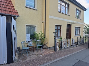 Un edificio residencial con fachada amarilla, un camino de entrada pavimentado, un pequeño espacio al aire libre con mesa y sillas, y una fila de esquíes apoyados en una valla.