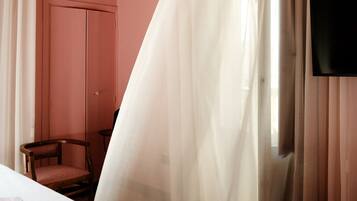 Habitación de hotel con cama, una silla, un televisor y ventana con cortinas traslúcidas.