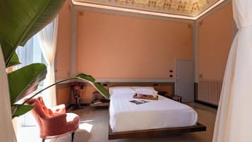 Quarto de hotel com uma cama grande, cabeceira de madeira, mesa de cabeceira com abajur, uma cadeira e uma janela ampla com cortinas.
