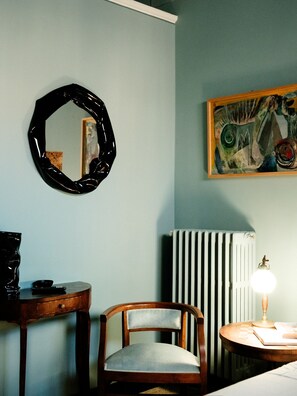 Una habitación con una silla de madera, un espejo redondo, un radiador y una mesa con una lámpara.