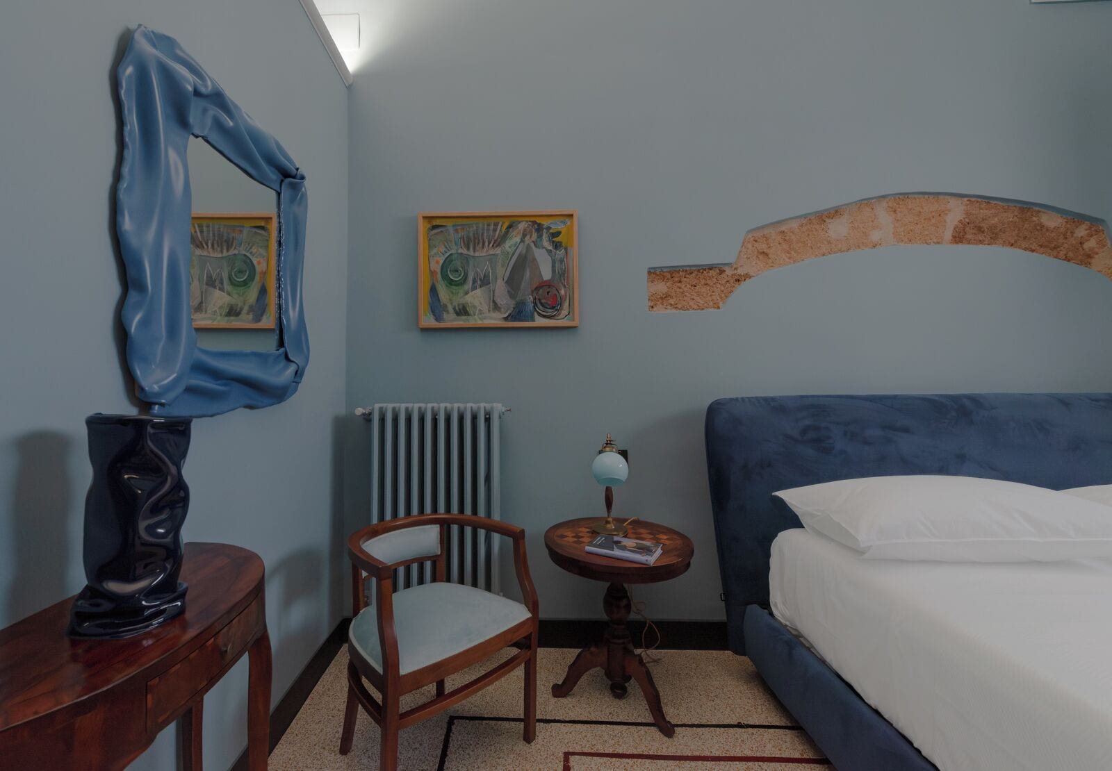 A bedroom with a bed, a chair, a table with a lamp, and two paintings on the wall.