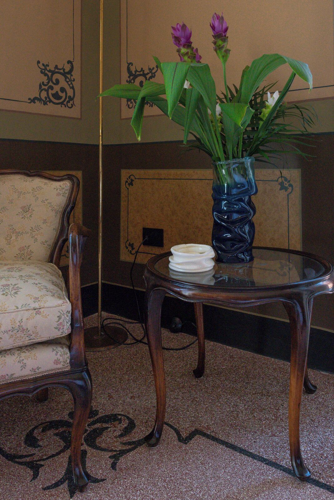 Una habitación con un sillón estampado con motivos florales, una mesa de vidrio con un jarrón de flores y una chimenea al fondo.