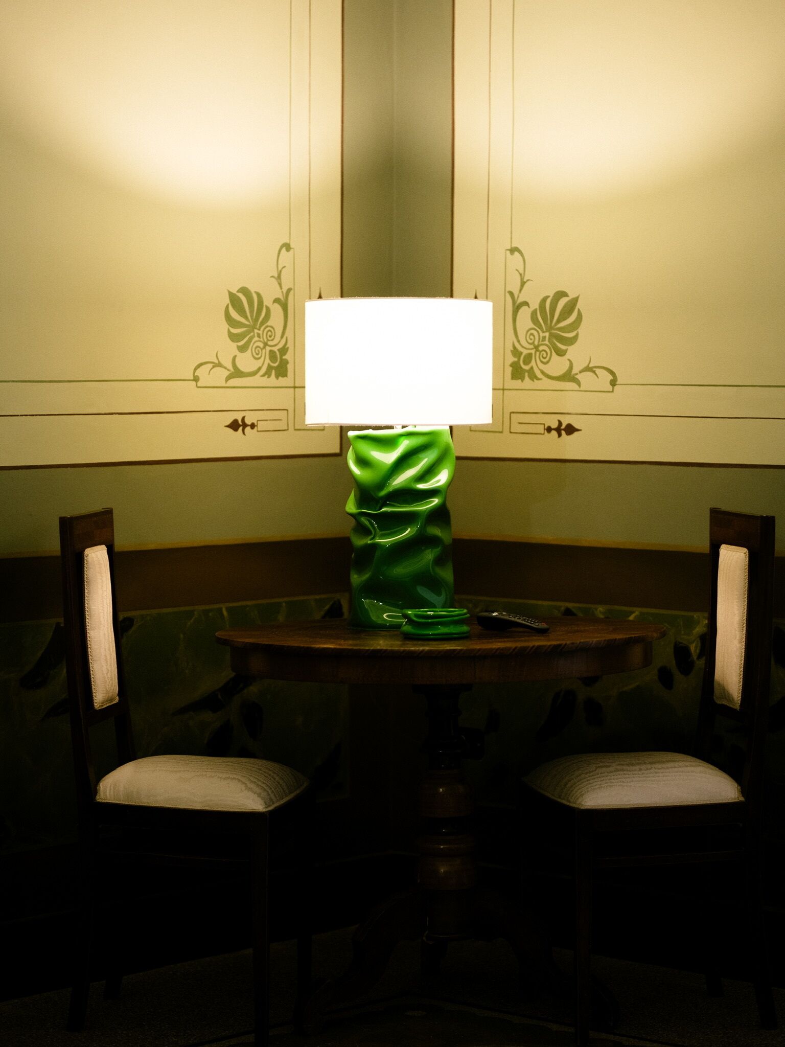 Una mesa con una lámpara verde, dos sillas y una pared con molduras decorativas.