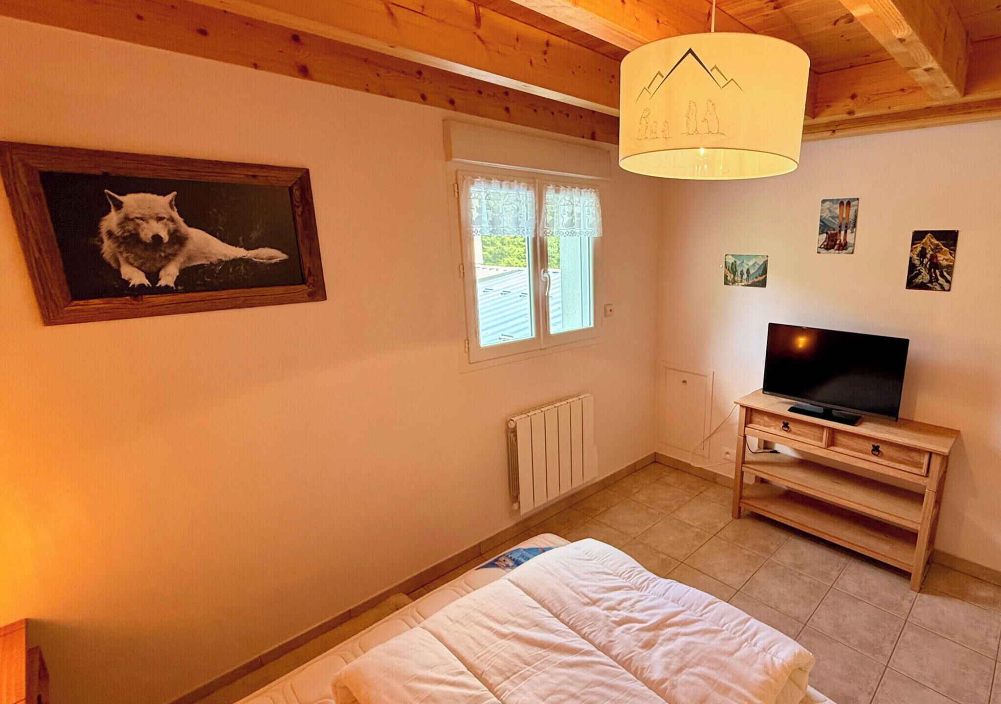 A bedroom with a bed, a television on a wooden stand, a painting of a dog, a window with curtains, and a radiator.