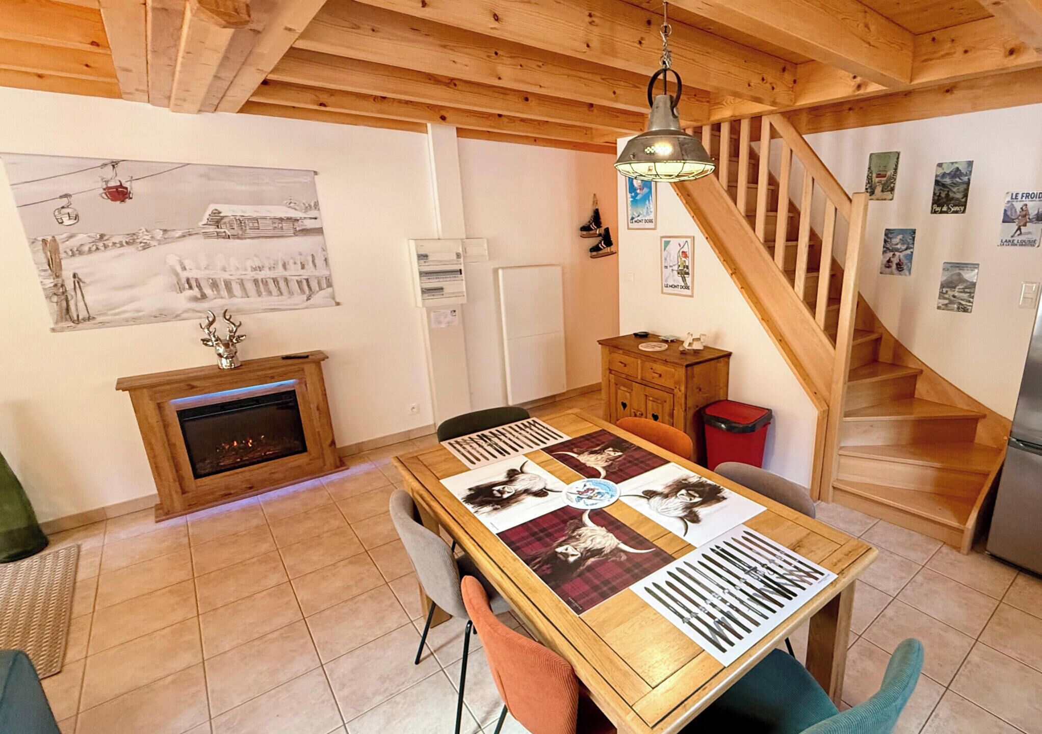Una habitación acogedora con chimenea, una mesa de comedor de madera con manteles individuales, una escalera y un refrigerador.