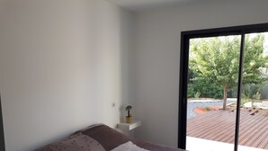 Une chambre à coucher avec un lit, une petite table de chevet, une plante en pot et une vue sur une terrasse extérieure, visible à travers une grande fenêtre.
