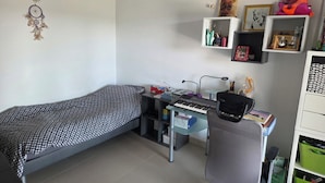 A bedroom with a single bed, a desk with a keyboard, a chair, and shelves with various items.