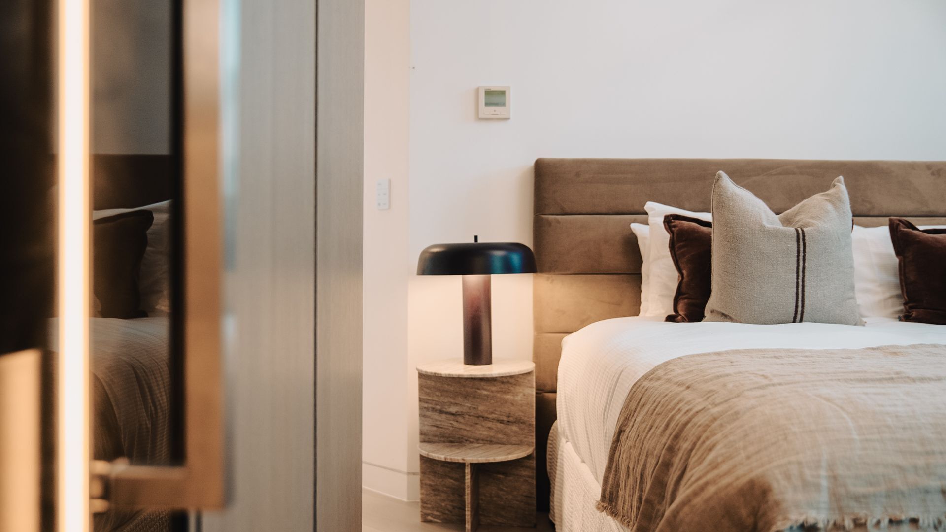 Une chambre d’hôtel équipée d’un lit, d’une table de chevet, d’une lampe et d’un thermostat fixé au mur.