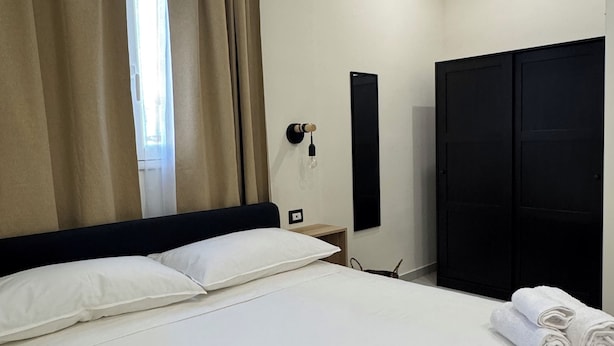 Une chambre d’hôtel avec un lit, des oreillers blancs, une armoire noire et une fenêtre avec des rideaux.