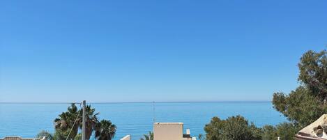 A balcony with yellow chairs and a table, overlooking a clear blue sea and a cityscape.