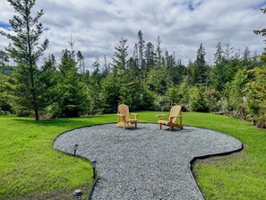 A circular outdoor seating area with two wooden Adirondack chairs and a table, surrounded by a gravel path and lush greenery.