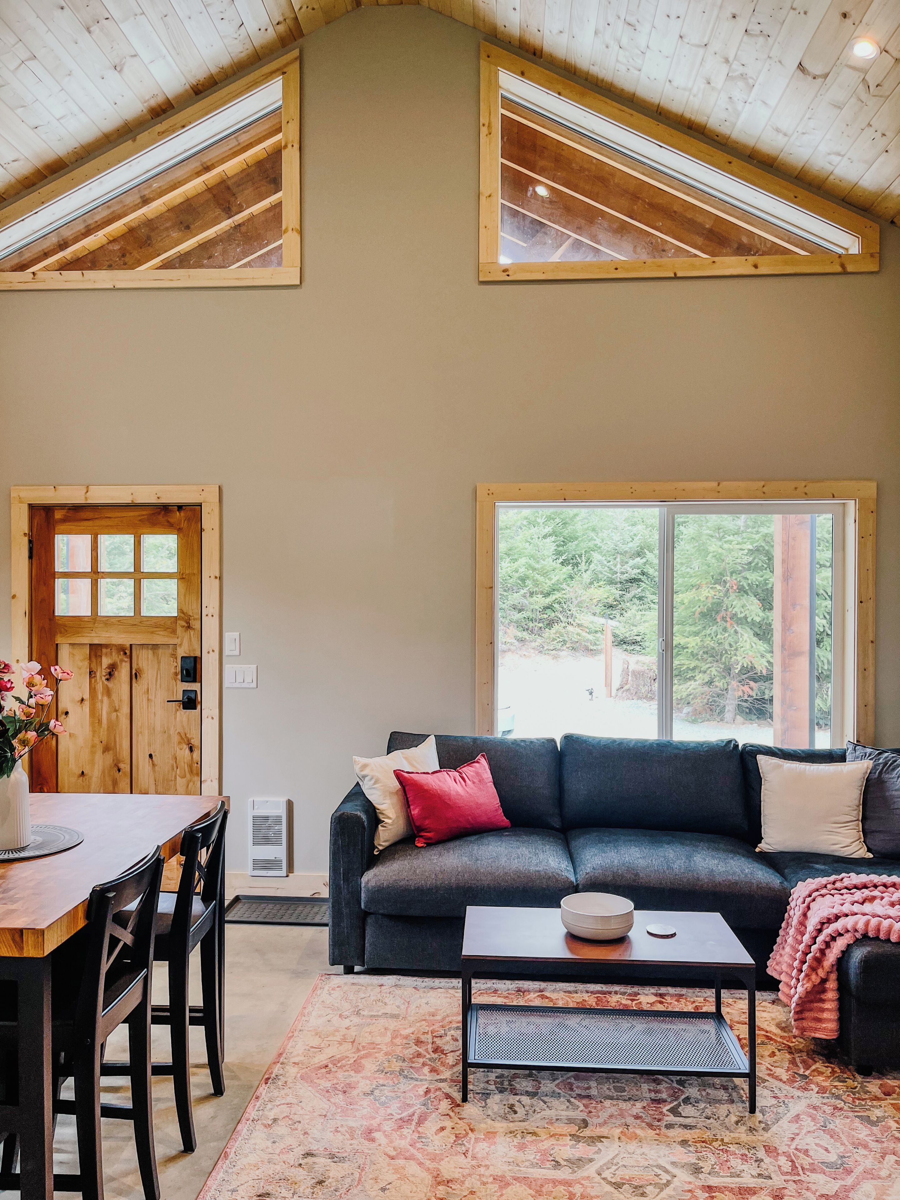 A room with a high ceiling, a dark-colored sofa, a coffee table, a dining table with chairs, and a large window with a view of greenery.