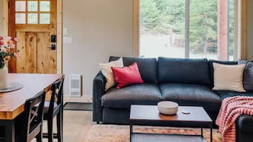 A room with a high ceiling, a dark-colored sofa, a coffee table, a dining table with chairs, and a large window with a view of greenery.