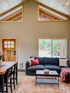 Une pièce avec un plafond haut, un canapé de couleur sombre, une table basse, une table à manger avec des chaises et une grande fenêtre donnant sur un paysage verdoyant.