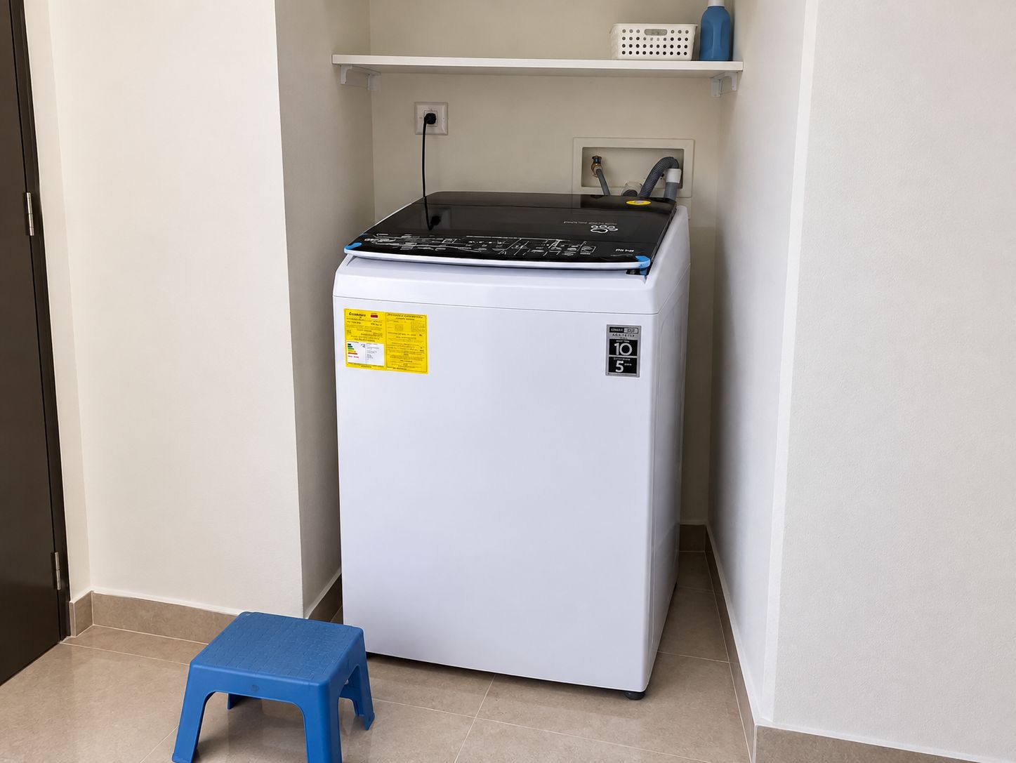 A compact refrigerator in a corner with a blue stool beside it.