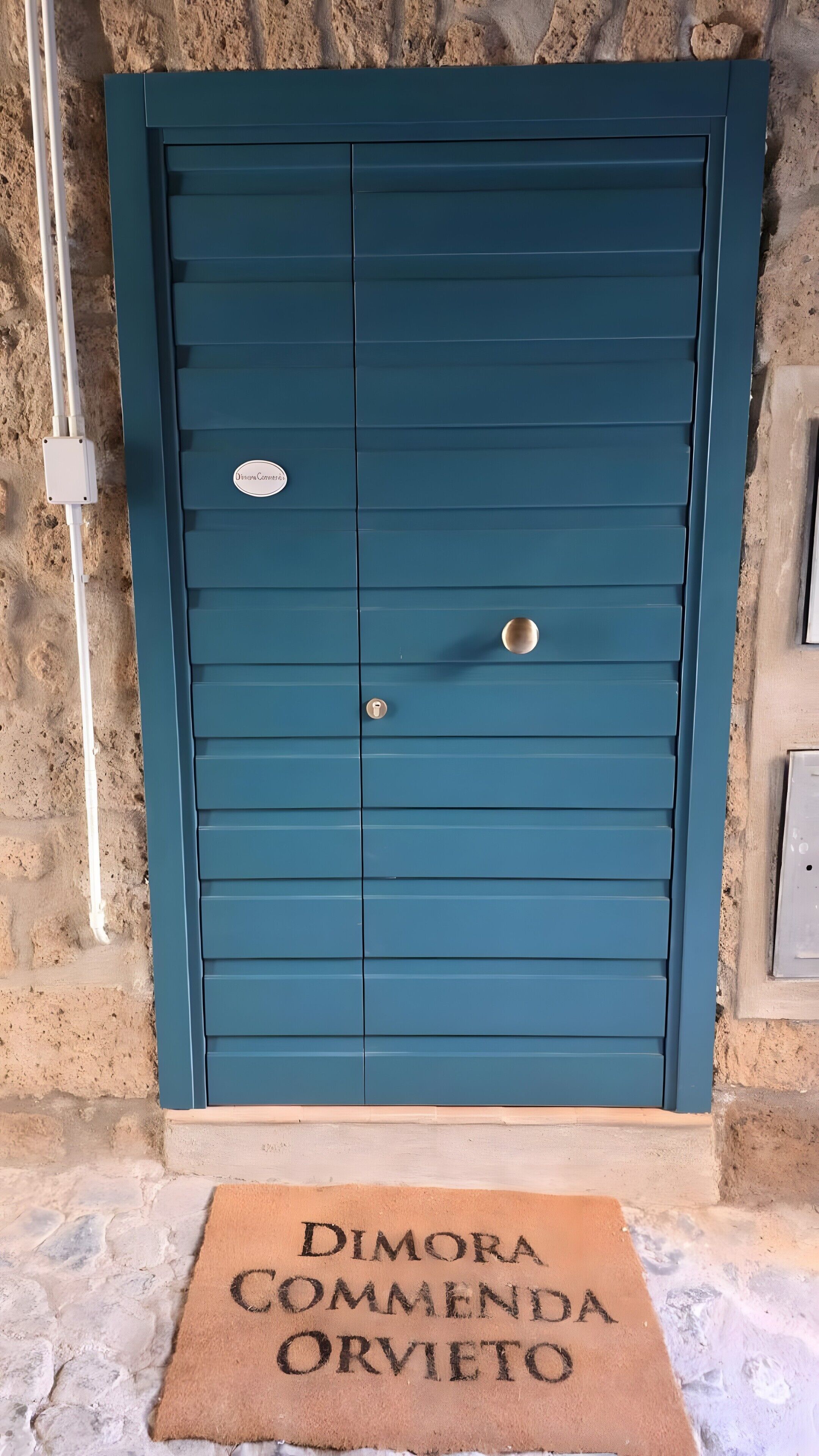 Una puerta color turquesa con un ojo de cerradura y un cerrojo, un felpudo con la inscripción 'Dimora Commenda Orvieto', y una pared de piedra.