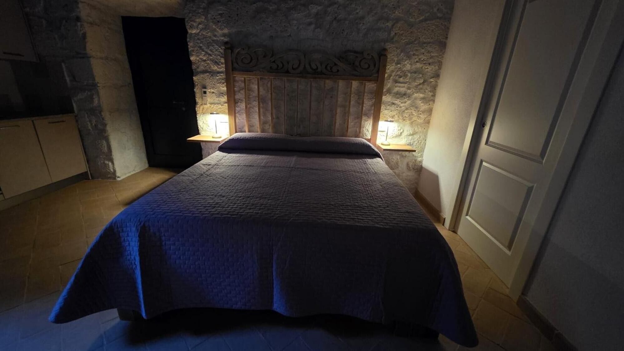 Ein Schlafzimmer mit Steinwand, einem Holz-Kopfteil, einem Bett mit blauer Bettdecke, zwei Nachttischlampen und einem Nachttisch aus Holz.