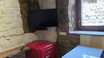 A small room with a stone wall, a red appliance, a flat-screen TV mounted on the wall, and a window with a view of a building outside.
