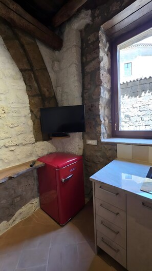 Una habitación pequeña con pared de piedra, un electrodoméstico rojo, un televisor de pantalla plana montado en la pared y una ventana con vista a un edificio.