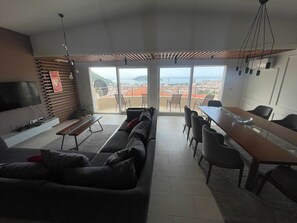 Ein modernes Wohnzimmer mit einer Couch, einem Esstisch und Stühlen, einem Fernseher und einem großen Fenster mit Blick auf Stadt und Berge.