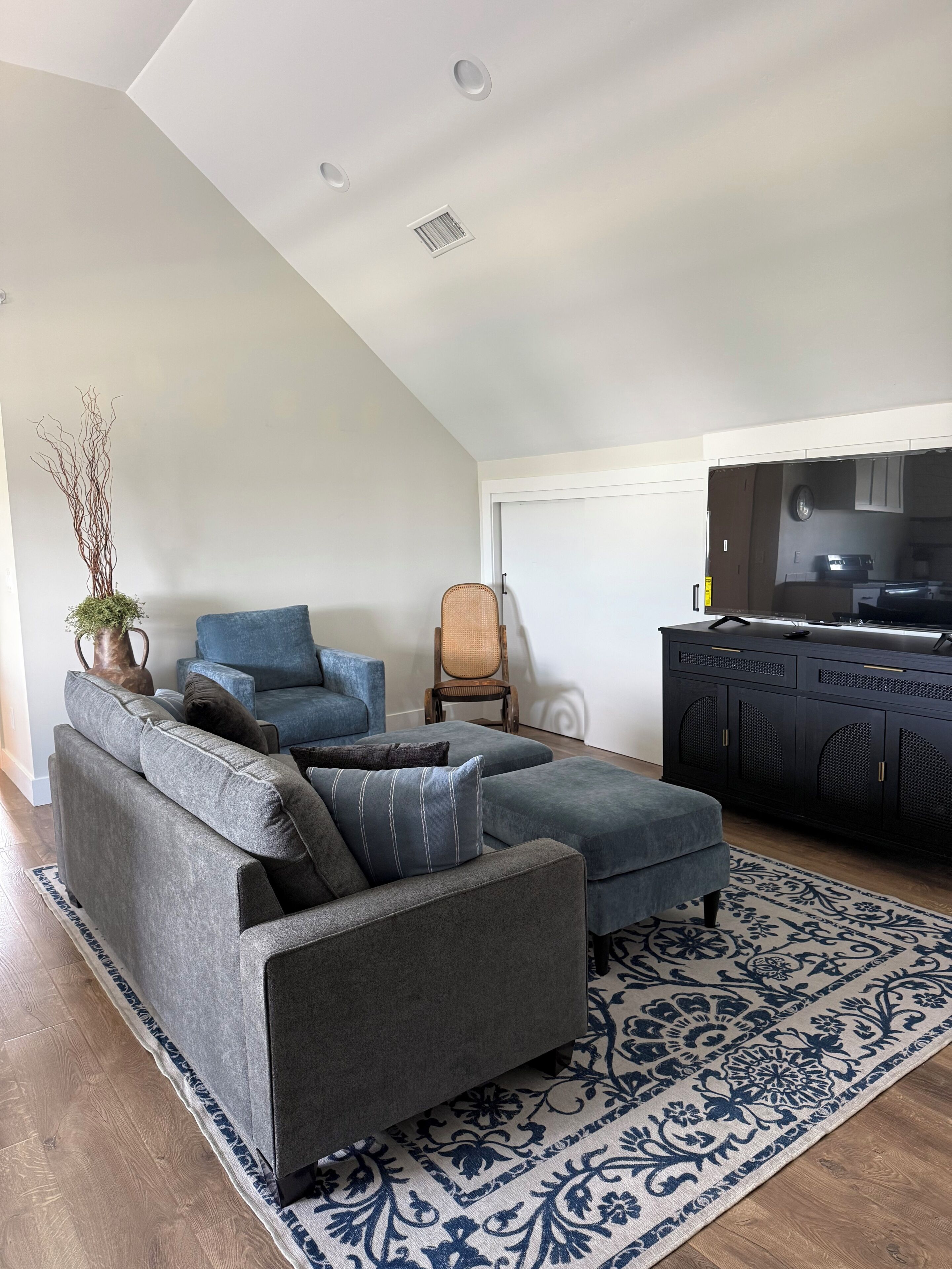 A modern living room with a gray sectional sofa, a blue armchair, a wooden chair, a black entertainment unit, and a large flat-screen TV.