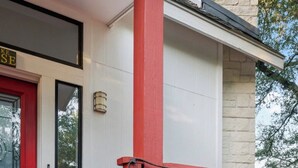 Eine rote Holztret Treppe führt zu einer weißen Tür mit schwarzem Rahmen, ein blauer Pflanzkasten befindet sich auf der Veranda, und im Hintergrund ist eine Steinmauer zu sehen.