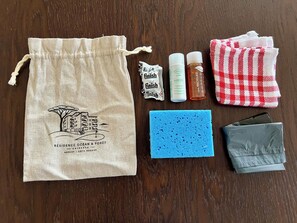 Uma sacola com cordão, ilustração de um edifício, duas garrafas de produto de limpeza para pisos, uma esponja azul, um pano xadrez vermelho e branco, e uma carteira cinza.
