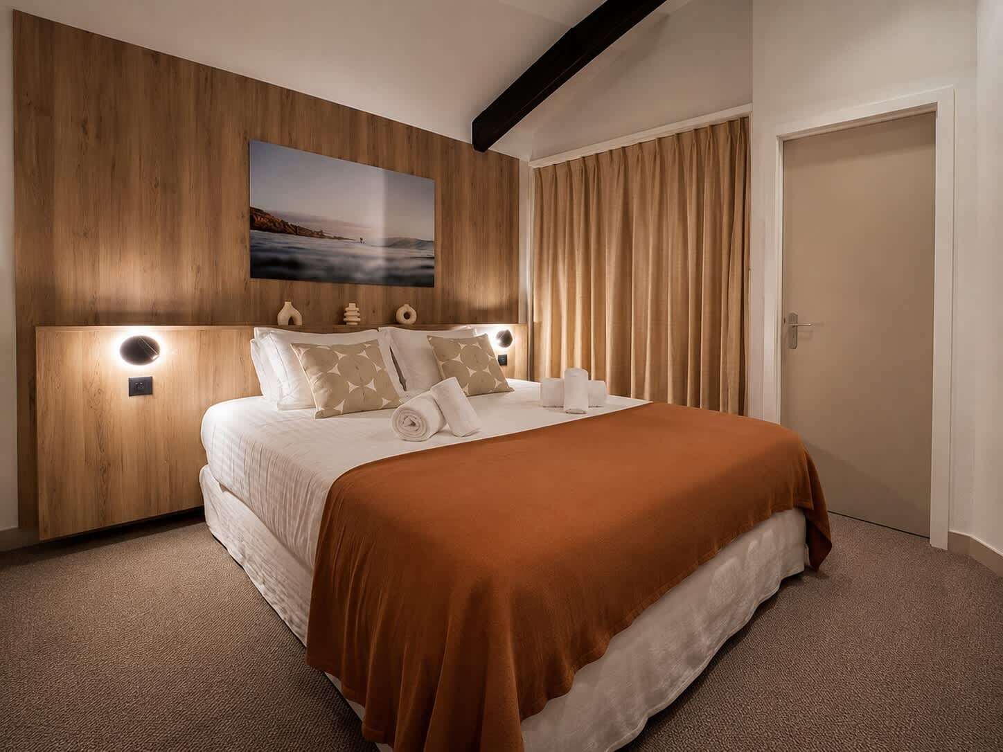 A modern hotel room with a wooden headboard, a large bed with a brown bedspread, and a painting above the bed.