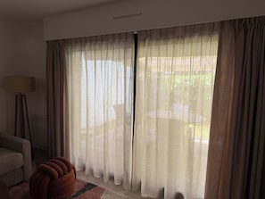 A room with a large window covered by sheer curtains, a floor lamp, a striped sofa, and a patterned rug. - T2 appartement 1 chambre, en RDJ avec terrasse (Anglet)