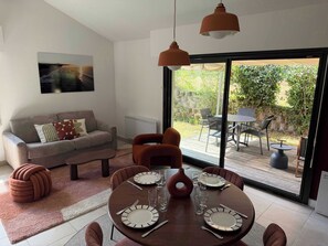 A modern living room with a dining area, a sofa, a coffee table, and a view of the outdoors through large windows. - Residence Océan & Forêt : T2 appartement 1 chambre, en RDJ avec terrasse (Anglet)