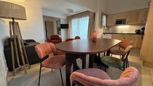 A modern living room with a round table, six chairs, a sofa, a lamp, and a kitchen area with a microwave. - T3 appartement 2 chambres RDJ avec terrasse (Anglet)