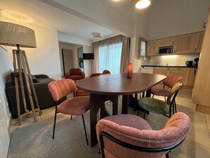 A modern living room with a round table, six chairs, a sofa, a lamp, and a kitchen area with a microwave. - T3 appartement 2 chambres RDJ avec terrasse (Anglet)