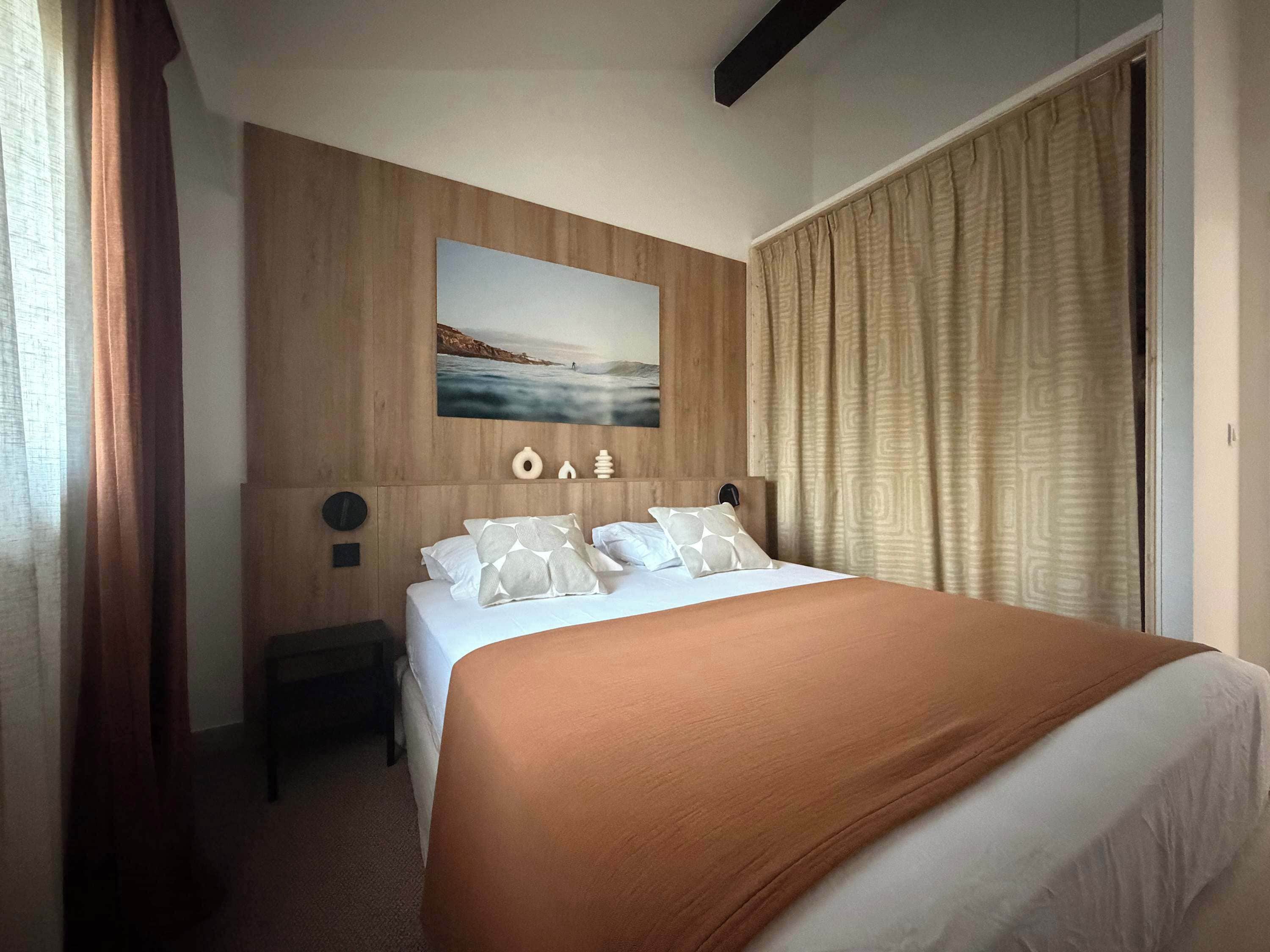 A hotel room with a bed, a nightstand, a wall-mounted picture, and a window with curtains.