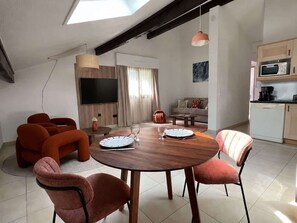 Ein modernes Wohnzimmer mit einem runden Holztisch, orangefarbenen Sofas, einem Flachbildfernseher und einer Küchenzeile mit einer Mikrowelle.