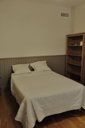 Een enkel bed met een witte spreidek, een houten kast en een ventilatie rooster aan de muur.