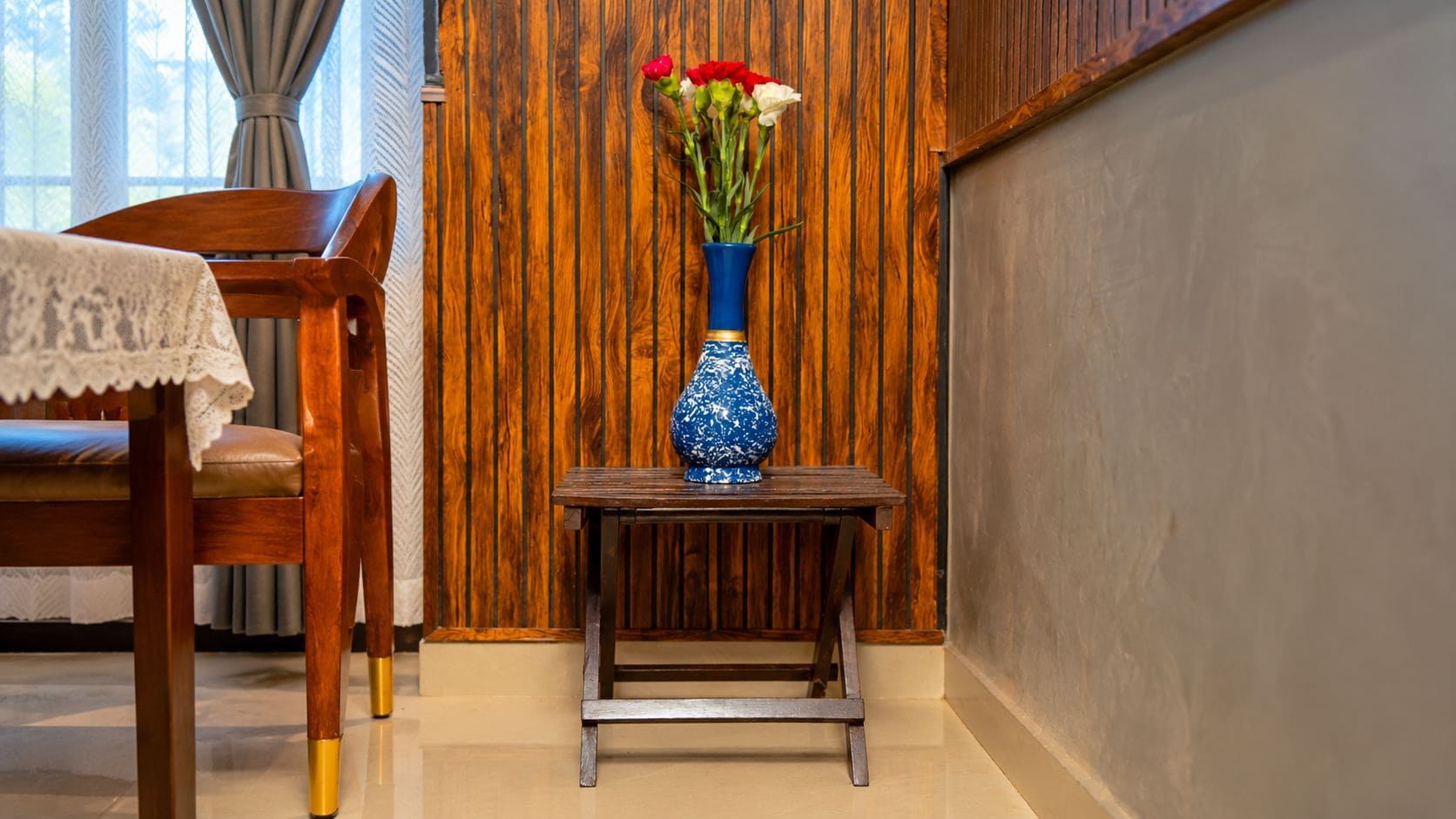 A wooden chair with a lace tablecloth, a small wooden table with a vase of flowers, and a wooden wall with vertical planks.
