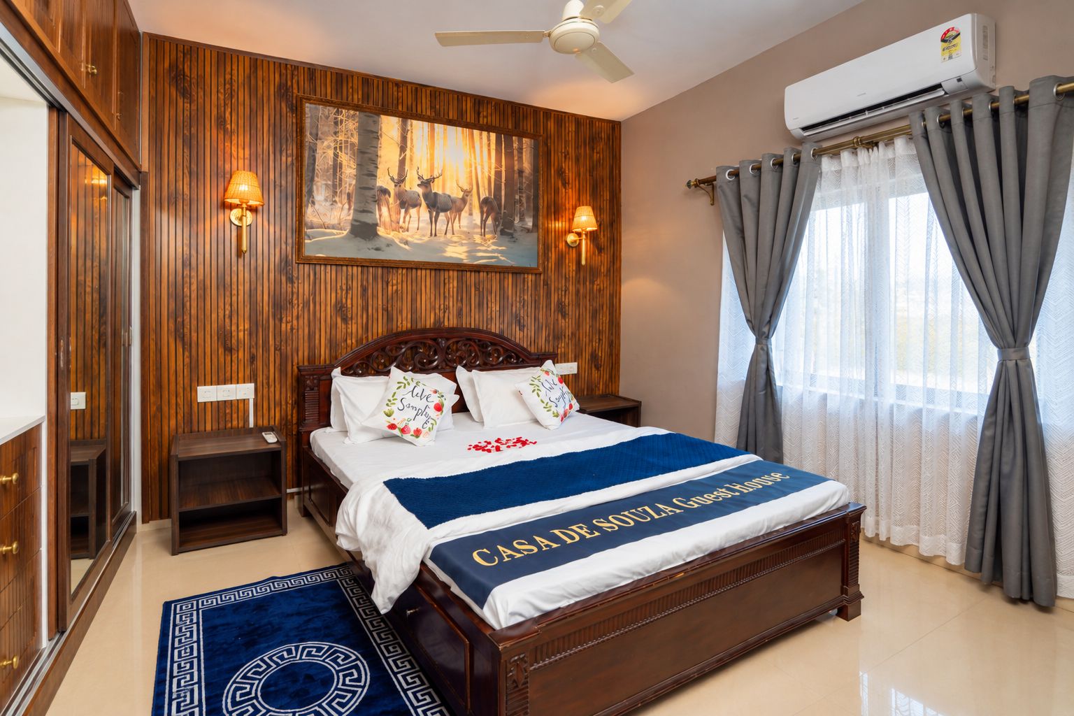 A hotel room with a wooden headboard, a bed with a blue and white bedspread, a nightstand, a wall-mounted lamp, and a large window with grey curtains.