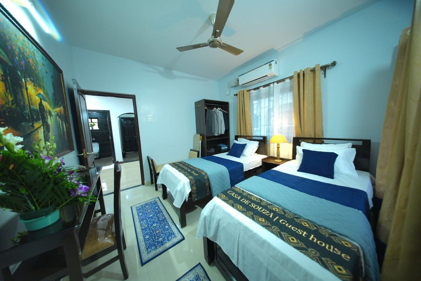 A hotel room with two beds, a ceiling fan, an air conditioning unit, a window with curtains, a painting on the wall, and a potted plant.